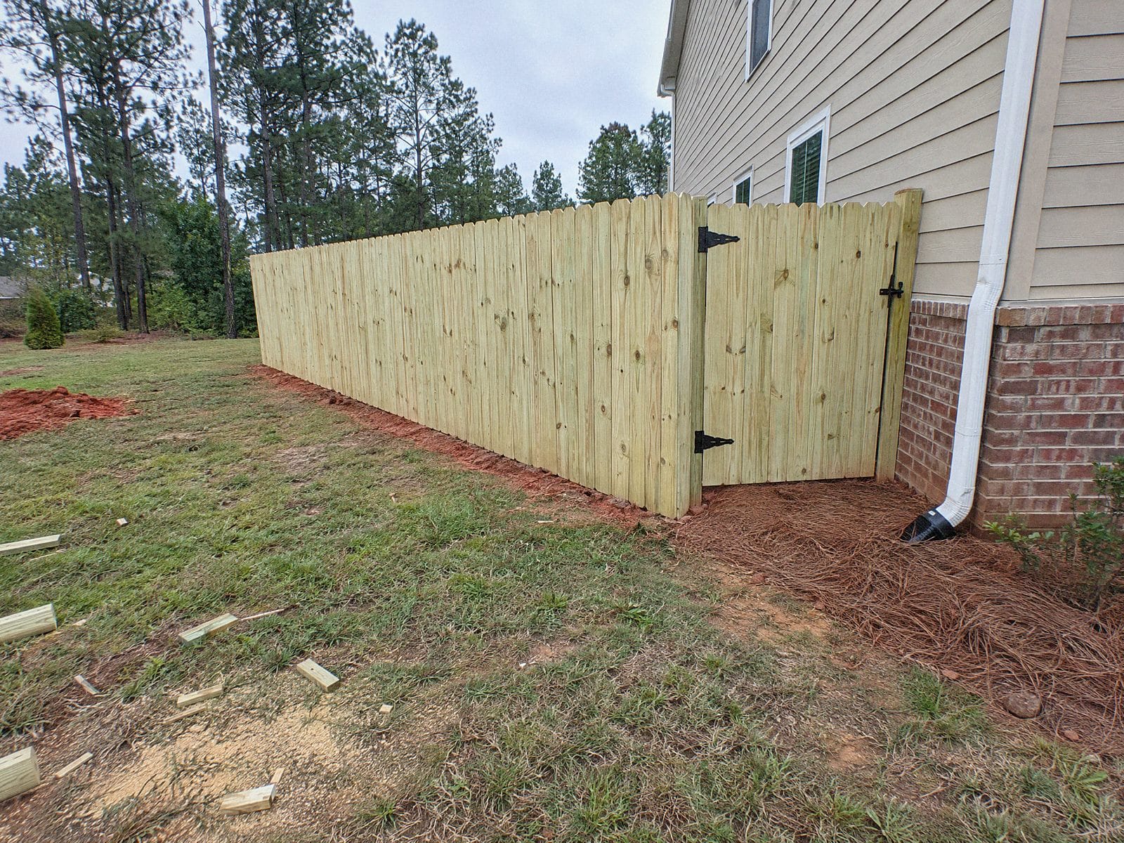 Six foot wood privacy fence meeting the back corner of a house, with a matching walk gate on black hinges and a black latch